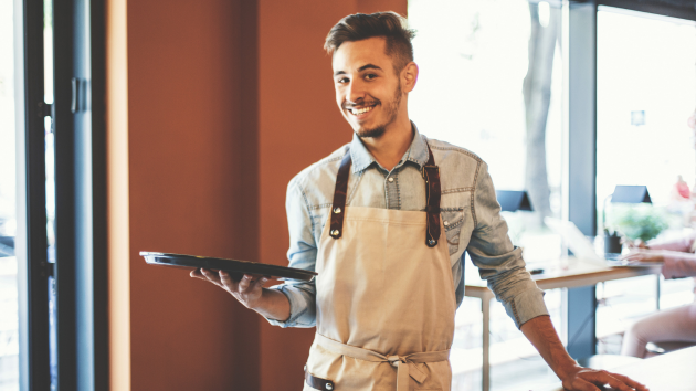 Lächelnder Kellner mit Schürze hält ein Tablett im Café.