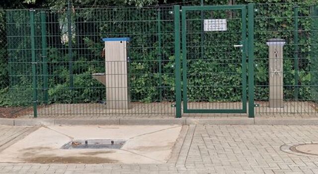 Grün eingezäunter Bereich mit Tor und Säulen für die Ver- und Entsorgung auf Pflastersteinen.