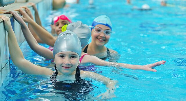 Kinder mit Badekappen schwimmen und halten sich am Beckenrand fest.