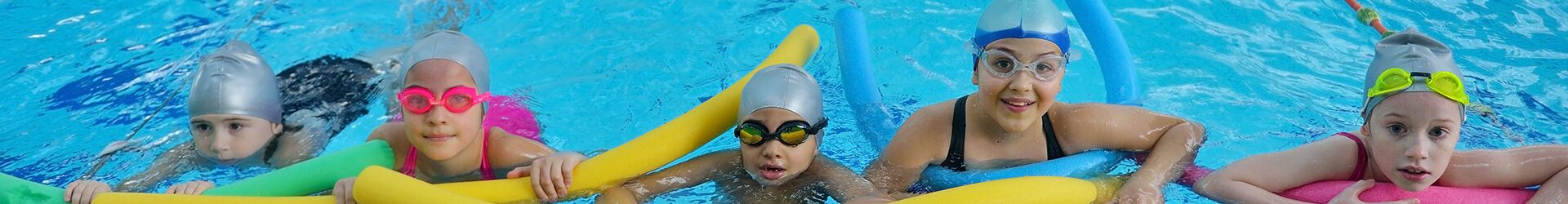 Kinder schwimmen mit Poolnudeln und Schwimmbrillen im Schwimmbecken.