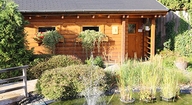 Blockhaussauna mit Blumen, Teich und Garten im Vordergrund.