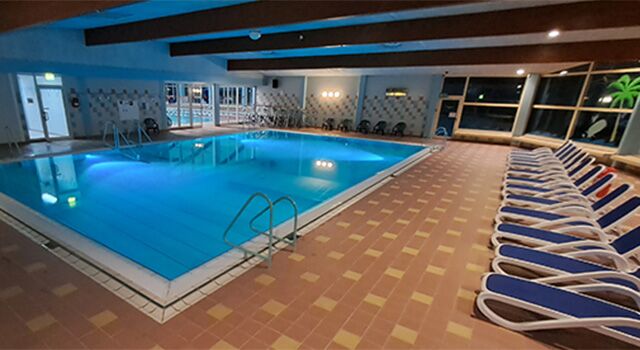 Leeres Hallenbad mit Liegestühlen und beleuchtetem Pool.