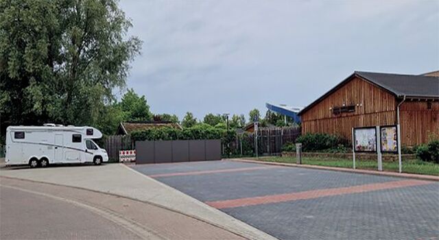 Wohnmobil auf einem Wohnmobilstellplatz neben einem Holzhäuser bei bewölktem Himmel.